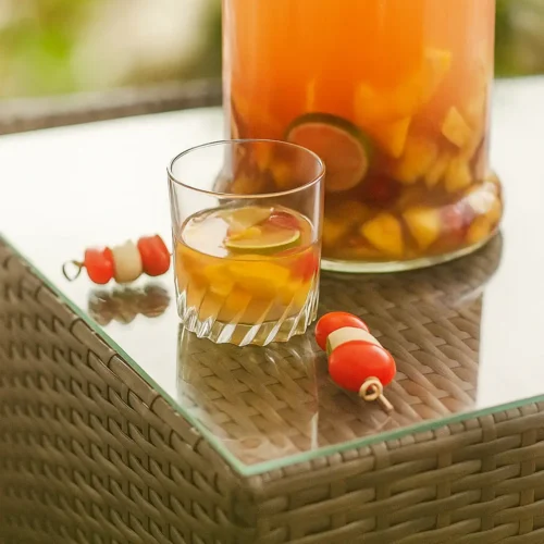 Verre de sangria rosée avec des morceaux de fruits frais — pêche, citron vert et orange — posé sur une table de jardin en rotin, à côté d’une grande carafe de sangria et de petites brochettes apéritives tomates-mozzarella, dans une ambiance estivale et ensoleillée.